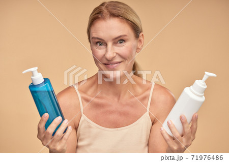 Portrait of a beautiful mature woman choosing between two cosmetic products while standing isolated over beige background in studio 71976486