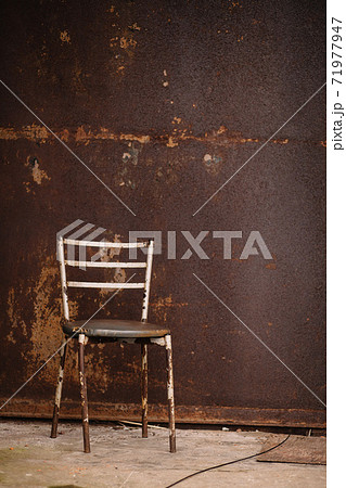 A rusty chair with peeling paint and a shabby leatherette seat against a rusty wall on the concrete floor. 71977947