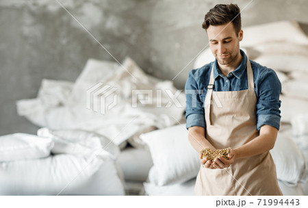 Smiling millennial brewery owner or worker in apron holds wheat or barley grains in hands in warehouse Smiling millennial brewery owner or worker in apron holds wheat or barley grains in hands in warehouse 71994443