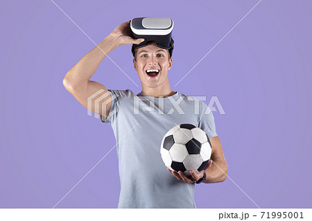 Exercising with virtual reality concept. Young athletic guy in VR headset holding soccer ball on lilac studio background 71995001