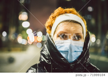 Portrait mature Caucasian woman in protective medical mask on face against background of evening city in winter. An elderly woman outdoors in cold weather during the coronavirus and covid 19 epidemic 71999104