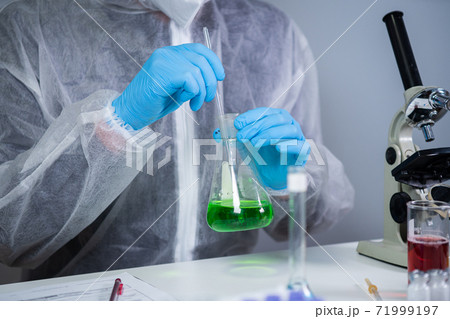 Technician in protective clothing hand stirring and shaking green liquid in Erlenmeyer flask in chemical laboratory. Laboratory technician studies chemical reactions and searches for cancer drug Technician in protective clothing hand stirring and shaking green liquid in Erlenmeyer flask in chemical laboratory. Laboratory technician studies chemical reactions and searches for cancer drug 71999197
