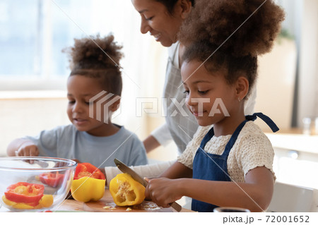 Smiling black foster mother watching adopted children cooking vegetable dish 72001652