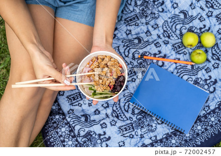 High angle of woman having picnic outdoor sitting on blue blanket with chopsticks eating takeout healthy vegetarian poke bowl with tofu, fermented soybeans -tempeh, cucumber, rice, carrots, onion High angle of woman having picnic outdoor sitting on blue blanket with chopsticks eating takeout healthy vegetarian poke bowl with tofu, fermented soybeans -tempeh, cucumber, rice, carrots, onion 72002457