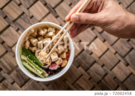Close up of a hand with chopsticks and healthy organic chicken poke bowl with cucumber, rice, carrots, onion served in takeout bowl on wooden table. Healthy lifestyle Close up of a hand with chopsticks and healthy organic chicken poke bowl with cucumber, rice, carrots, onion served in takeout bowl on wooden table. Healthy lifestyle 72002458
