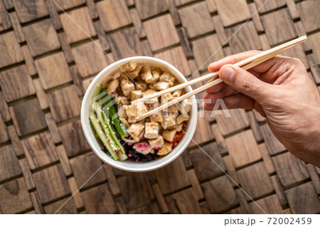 Close up of a hand with chopsticks and healthy organic chicken poke bowl with cucumber, rice, carrots, onion served in takeout bowl on wooden table. Healthy lifestyle Close up of a hand with chopsticks and healthy organic chicken poke bowl with cucumber, rice, carrots, onion served in takeout bowl on wooden table. Healthy lifestyle 72002459