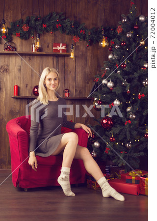 Beautiful young woman in a dress sitting in a red armchair in the New Year home interior near the Christmas tree. 72004012