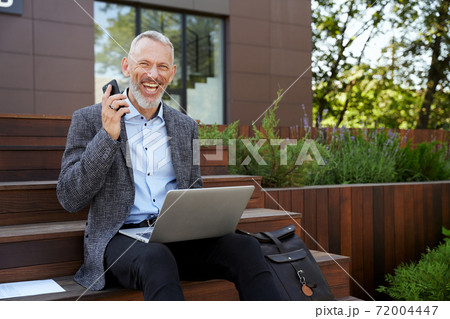 Improve your business. Joyful mature businessman smiling, making a call using his mobile phone while working on the laptop, sitting outdoors on the daytime Improve your business. Joyful mature businessman smiling, making a call using his mobile phone while working on the laptop, sitting outdoors on the daytime 72004447
