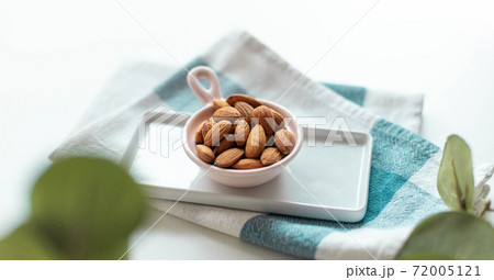 Fresh nut almonds in a ceramic bowl with towel on a white background. Green plant decor 72005121