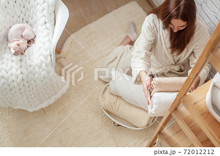 Woman in robe is sitting near wooden shelving, organizing closet by sorting and folding bath towels. Light pink colored Don Sphynx cat 72012212