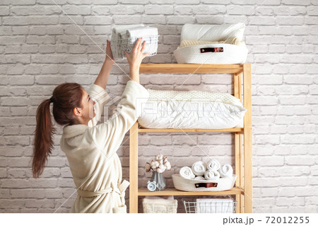 Young woman in warm robe is organizing linen closet with neatly folded towels, sheets and blankets. Young woman in warm robe is organizing linen closet with neatly folded towels, sheets and blankets. 72012255