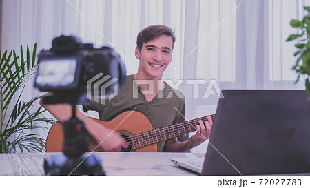 Teen recording his video blog. Teenager playing music in front of a video camera for his blog channel.. an blogger videotapes his vlog at home while playing guitar.  Blogger makes online streaming 72027783