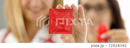 Doctors in white uniforms with red ribbon on chest hold condom in hand closeup Doctors in white uniforms with red ribbon on chest hold condom in hand closeup 72028392