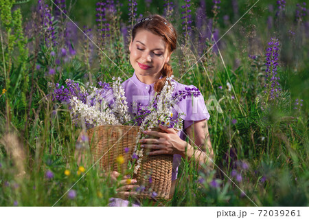 Portrait of young beautiful woman with bouquet of flowers in basket at sunny field. Pretty vintage girl in lilac dress and hat smiles at grassy forest meadow. 72039261