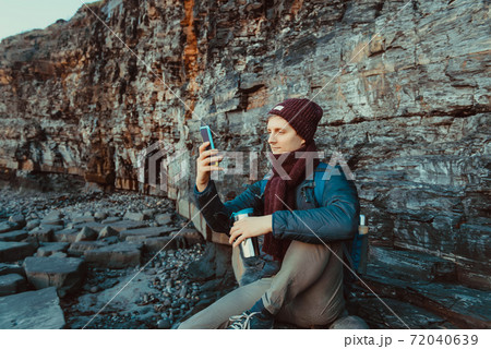 A young man in warm clothes sitting on the stone and taking pictures of the sunset on his cell phone camera. Rest during a walk on the rocky coast. Local self-traveling in autumn, winter. Copy space. A young man in warm clothes sitting on the stone and taking pictures of the sunset on his cell phone camera. Rest during a walk on the rocky coast. Local self-traveling in autumn, winter. Copy space. 72040639