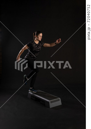 Fitness girl on a step platform, pulls her leg. Go in for sports on a black background Joyful Fitness girl on a step platform, pulls her leg. Go in for sports on a black background Joyful 72040752