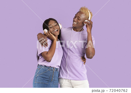 Happy black couple in headphones listening to music and hugging over lilac studio background Happy black couple in headphones listening to music and hugging over lilac studio background 72040762