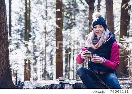 A smiling tourist is resting on a tree trunk, drinking hot tea and eating a hamburger on a mountain trail on a cold day. A smiling tourist is resting on a tree trunk, drinking hot tea and eating a hamburger on a mountain trail on a cold day. 72042111