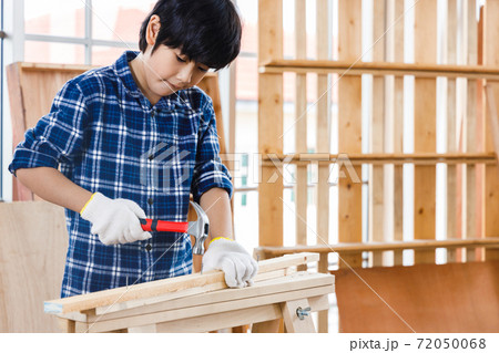 Asian boy nails timber wood using hammer in carpentry workshop. 72050068