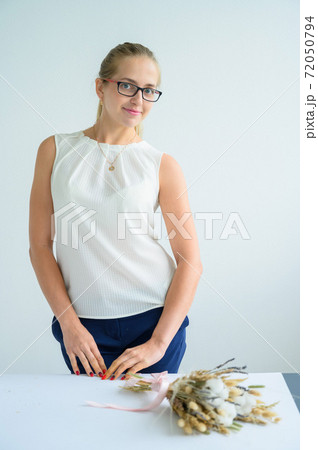 The blonde in glasses and a white blouse collects a bouquet of dried flowers and cotton in a white studio. A charming florist smiles at work. 72050794