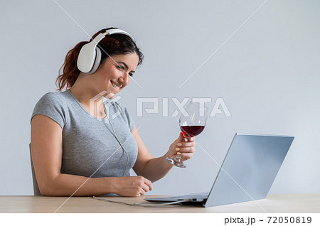 Caucasian woman in headphones toasting with a glass of red wine during a video call at home. Remote communication in isolation Caucasian woman in headphones toasting with a glass of red wine during a video call at home. Remote communication in isolation 72050819