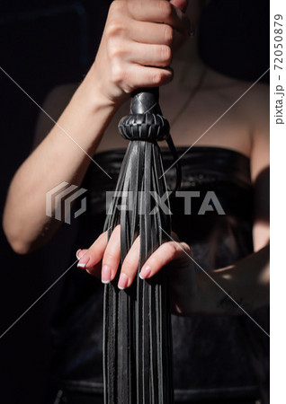 BDSM concept. A woman in a leather dress holds a black whip with a long fringe. Role-playing games for adults. Close-up of sex toys for domination. BDSM concept. A woman in a leather dress holds a black whip with a long fringe. Role-playing games for adults. Close-up of sex toys for domination. 72050879