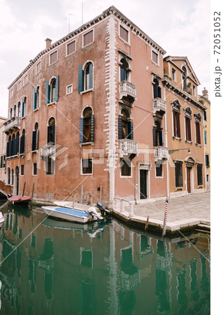 Wide angle shot of a three-story house, peach-colored, near a small narrow Venetian Canal. A motor boat moored near the walls of a house standing in the water in Venice, Italy. Wide angle shot of a three-story house, peach-colored, near a small narrow Venetian Canal. A motor boat moored near the walls of a house standing in the water in Venice, Italy. 72051052