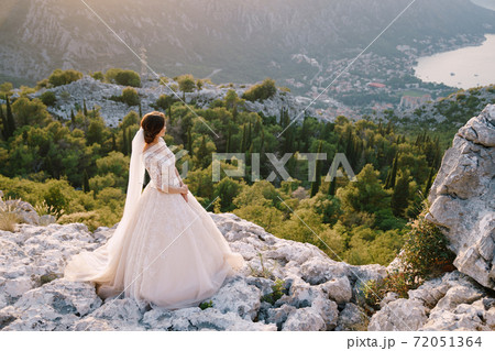The bride stands on top of a mountain with panoramic views of the Bay of Kotor, at sunset. Fine-art destination wedding photo in Montenegro, Mount Lovchen. 72051364