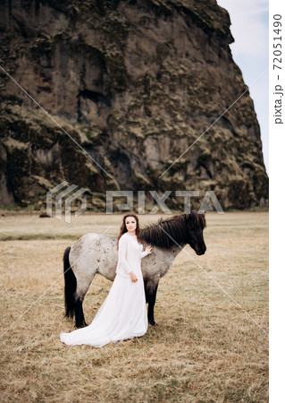 A girl in a white dress next to a white horse, with black ones. legs and mane. In a field of dry grass, against the backdrop of a cliff. Destination Iceland wedding photo session with Icelandic horses 72051490