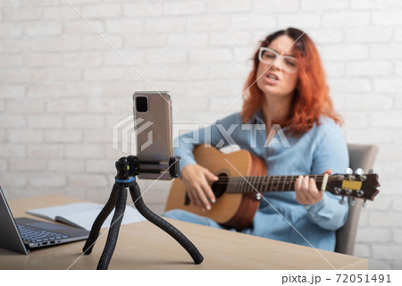 A woman is broadcasting online on her phone at home. The girl sings and plays the guitar live A woman is broadcasting online on her phone at home. The girl sings and plays the guitar live 72051491