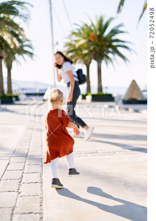 Little girl in a knitted terracotta dress is chasing her mother on a boat pier Little girl in a knitted terracotta dress is chasing her mother on a boat pier 72052498