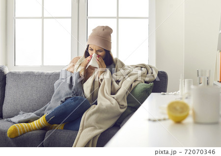 Young woman in warm clothing, socks and hat sitting on sofa, blowing nose and feeling ill Young woman in warm clothing, socks and hat sitting on sofa, blowing nose and feeling ill 72070346