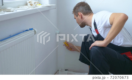 handsome man in work overalls paints the wall with a roller white paint handsome man in work overalls paints the wall with a roller white paint 72070673