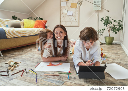 Family time. Teenage boy lying on the floor in living room, using digital tablet and doing homework online while his mother and sister playing together near him Family time. Teenage boy lying on the floor in living room, using digital tablet and doing homework online while his mother and sister playing together near him 72072903