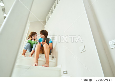 Homeschooling concept. Small brother and sister sitting on stairs at home using digital tablet and doing homework together Homeschooling concept. Small brother and sister sitting on stairs at home using digital tablet and doing homework together 72072931