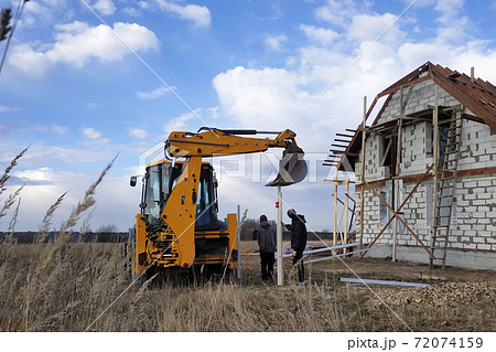 The excavator with a bucket pushes the pillars on the fence in the field near the house 72074159