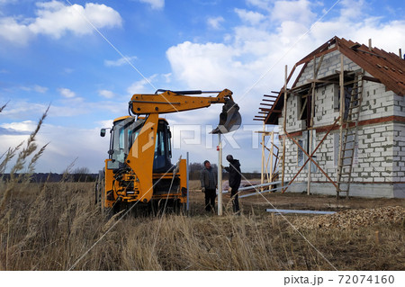 The excavator with a bucket pushes the pillars on the fence in the field near the house 72074160