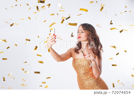 Young woman drinks champagne, makes a selfie on a smartphone, having fun with golden confetti 72074854