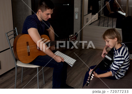 Young male musician teaches little boy student how to play the acoustic guitar and play by notes Young male musician teaches little boy student how to play the acoustic guitar and play by notes 72077233