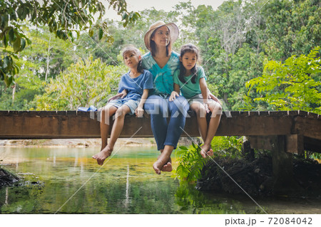 Two cute asian child girls and their mother sitting on the wooden bridge over the river and enjoy with nature together in summer time. 72084042