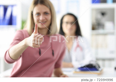 Patient holds thumb up in medical office in background doctor sits. 72094996