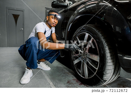 Professional African male worker in overalls and black protective gloves, cleaning alloy wheels rims of luxury car with a special brush for cast wheels in a detailing workshop, looking at camera 72112672