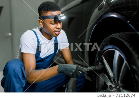 Car washing and detailing workshop. African man worker in protective overalls and rubber gloves, washing car alloy wheel on a car wash, using special brush. Close up portrait, selective focus Car washing and detailing workshop. African man worker in protective overalls and rubber gloves, washing car alloy wheel on a car wash, using special brush. Close up portrait, selective focus 72112674