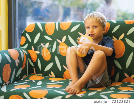 Front view of happy thirsty blond kid boy drinking lemonade, fruit tea, kombucha ice tea with peach and lemon, wellness orange drinks from plastic cup with straw, sitting on multi color sofa Front view of happy thirsty blond kid boy drinking lemonade, fruit tea, kombucha ice tea with peach and lemon, wellness orange drinks from plastic cup with straw, sitting on multi color sofa 72122822