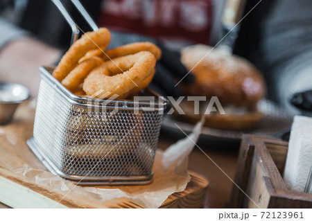 Breaded onion rings in square metal basket on wooden cutting board on table in restaurant. Burger and cream sauce on background. Fried onion rings, It is good appetizer crispy and testy. Breaded onion rings in square metal basket on wooden cutting board on table in restaurant. Burger and cream sauce on background. Fried onion rings, It is good appetizer crispy and testy. 72123961