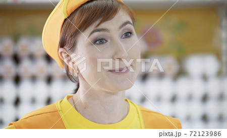 Portrait of smiling Caucasian female staff saleswoman talking to buyer and standing in grocery section of supermarket. Cheerful slender saleswoman in yellow t-shirt andorange apron. 72123986