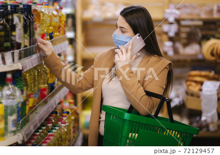 Buyer wearing a protective mask and gloves in supermarket. Shopping during the pandemic quarantine Covid-19. Nonperishable smart purchased household pantry groceries preparation. Woman spear by phone Buyer wearing a protective mask and gloves in supermarket. Shopping during the pandemic quarantine Covid-19. Nonperishable smart purchased household pantry groceries preparation. Woman spear by phone 72129457