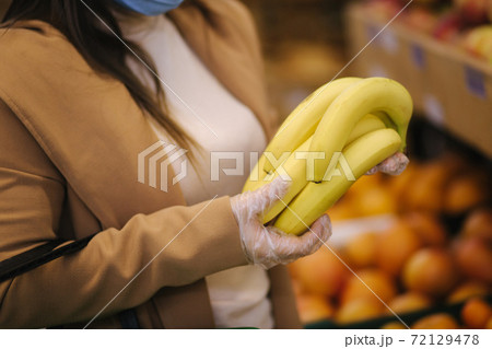 Close up of Young woman in protective gloves and face mask hold beautiful fresh bananas in hand. Beautiful young girl with food basket choosing food by stand with fruits. Shopping during quarantine 72129478