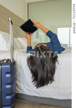 Woman lying on bed using tablet next to carry-on luggage 72129704
