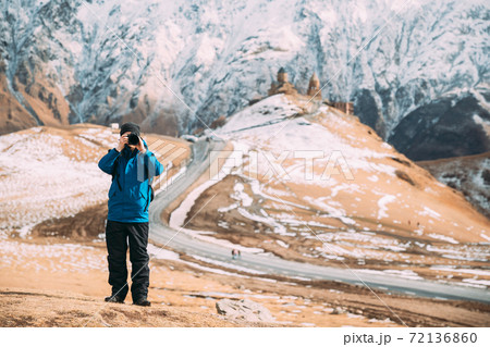 Stepantsminda, Gergeti, Georgia. Man Tourist Backpacker Traveler Photographer Taking Pictures Photos Of Holy Trinity Church - Tsminda Sameba. Beautiful Georgian Landscape In Early Winter Stepantsminda, Gergeti, Georgia. Man Tourist Backpacker Traveler Photographer Taking Pictures Photos Of Holy Trinity Church - Tsminda Sameba. Beautiful Georgian Landscape In Early Winter 72136860
